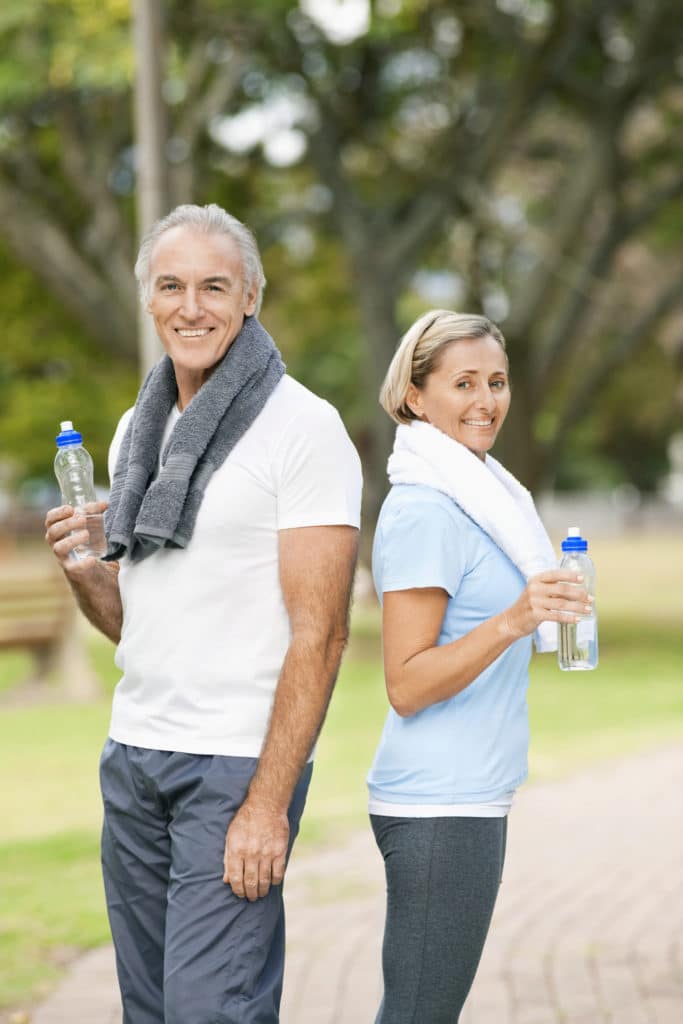 Portrait of a senior couple after exercise. They are standing back to back with towels draped around their necks and are holding bottles of water. Both are smiling at the camera. Vertical shot.