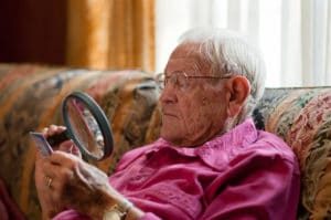 Elderly old man using magnifying glass to read fine print on object with strong light over shoulder while wearing glasses.