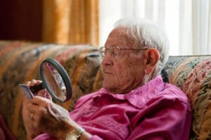 Elderly old man using magnifying glass to read fine print on object with strong light over shoulder while wearing glasses.