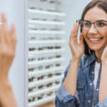 Smiling woman choosing eyeglasses and looking at mirror in optical shop