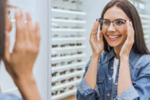 Smiling woman choosing eyeglasses and looking at mirror in optical shop